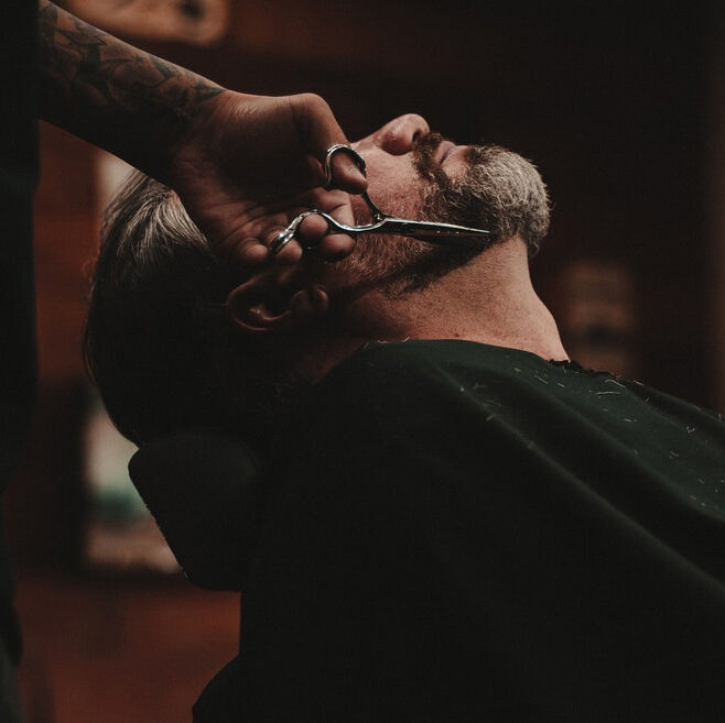 Man getting his beard trimmed with scissors by a barber in a dimly lit setting.